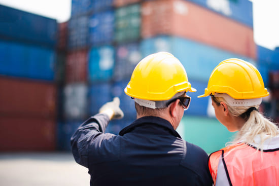 Zwei Arbeiter in Sicherheitskleidung stehen vor gestapelten Containern in einem Hafen